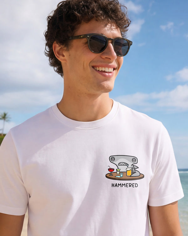 Man wearing a white t-shirt with the Hammered Shark logo and olive cargo shorts standing on a beach. Close up