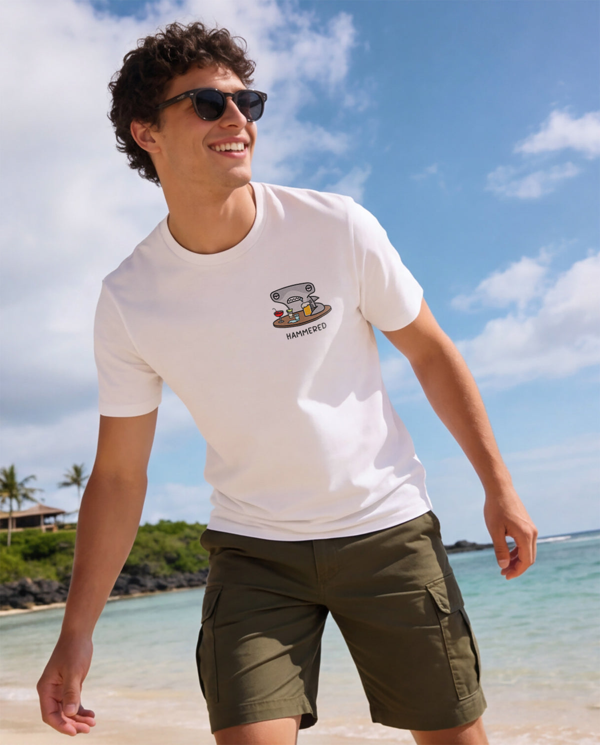 Man wearing a white t-shirt with the Hammered Shark logo and olive cargo shorts standing on a beach.