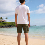 Man wearing a white t-shirt with the Hammered Shark logo and olive cargo shorts standing on a beach. Back view