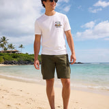 Man wearing a white t-shirt with the Hammered Shark logo and olive cargo shorts standing on a beach.