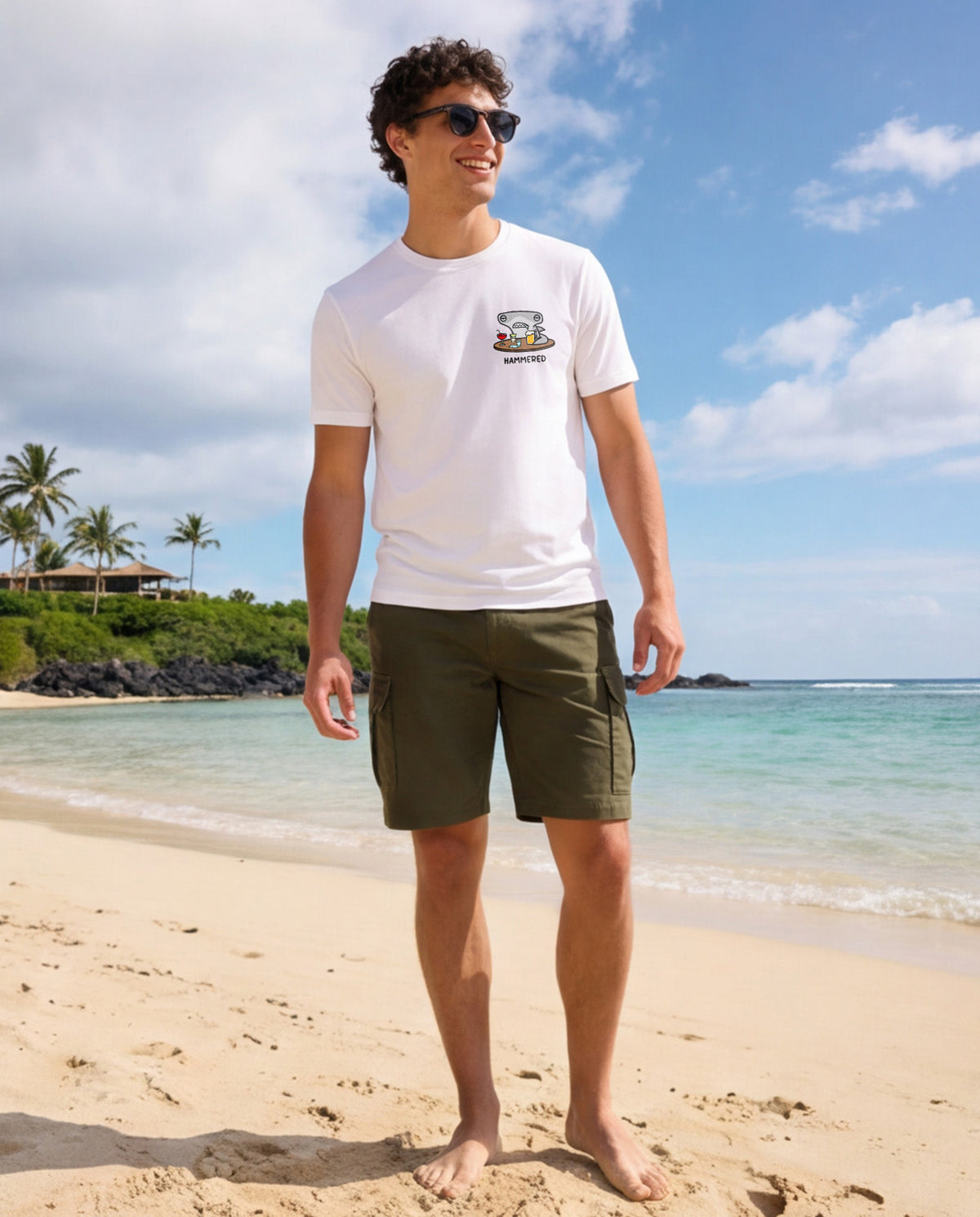 Man wearing a white t-shirt with the Hammered Shark logo and olive cargo shorts standing on a beach.